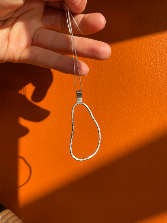 A hand holds a silver pendant necklace with a textured, hammered finish against a vibrant orange wall with strong sunlight and shadows.