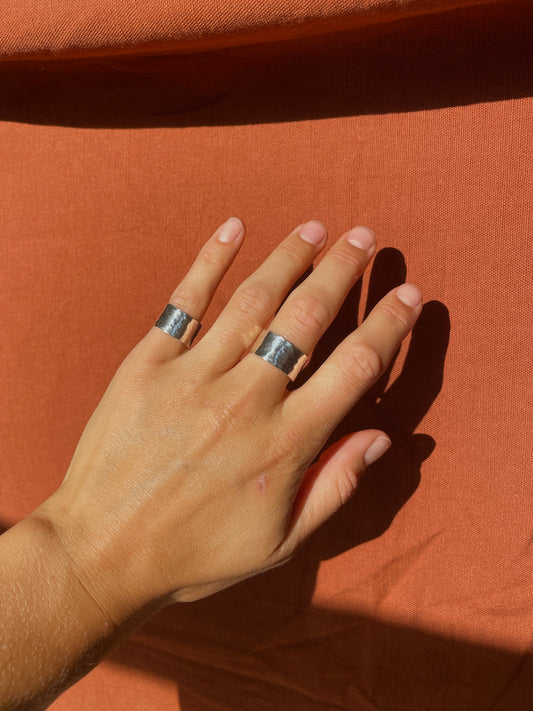 A close-up of a hand wearing two wide silver rings with textured, hammered surfaces, resting against a draped terracotta fabric background.