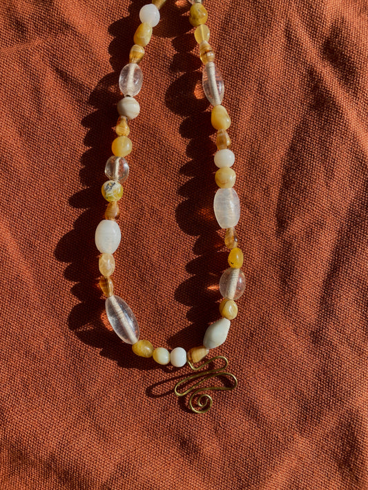 A beaded necklace with translucent white beads, lemon opal beads, and clear glass beads, featuring a small brass spiral charm, laid on a textured rust-orange fabric background.
