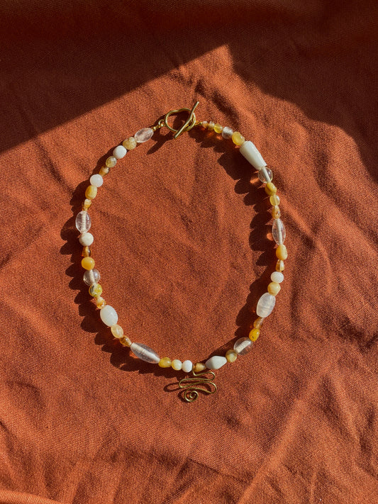 A beaded necklace with white, opal, and clear beads, featuring a gold toggle clasp and a small brass script charm, displayed on a textured rust-orange fabric background.
