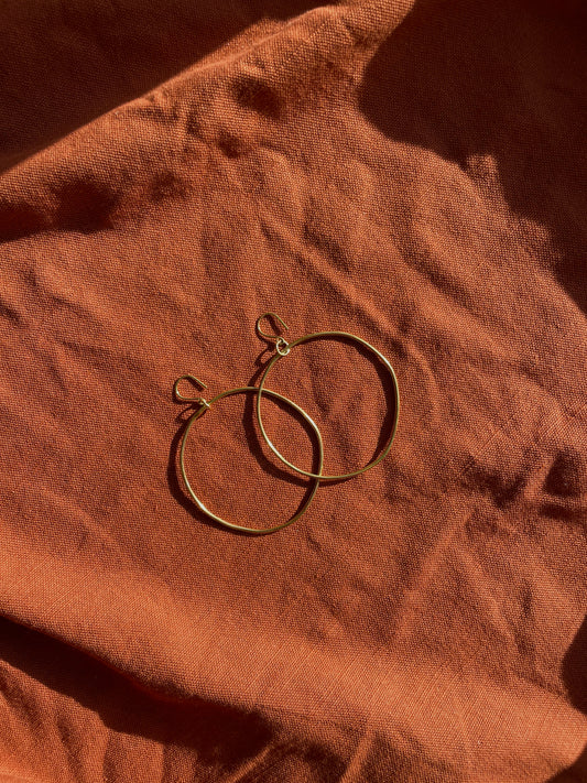 Two thin brass hoop earrings resting on a textured terracotta fabric background under warm directional sunlight.