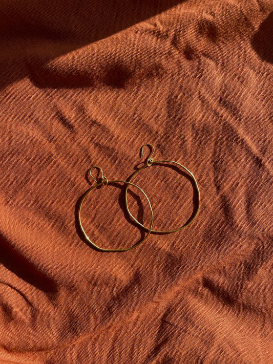Two thin brass hoop earrings with fishhook backs, resting on a textured, draped terracotta fabric under warm directional sunlight.